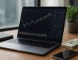 A laptop displaying trading charts on a wooden desk alongside a notebook, a smartphone, and some cash